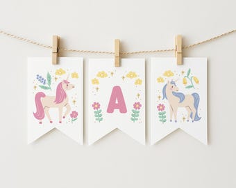 May include: Three white pennant banners with unicorn and floral designs, suspended by wooden clothespins. The banners feature a pink unicorn, the letter "A", and a unicorn with blue hair. The banners are decorated with flowers and stars.