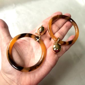 May include: A pair of large, round, brown and black tortoiseshell clip-on earrings with gold accents. Each earring has a dangling gold ball charm.