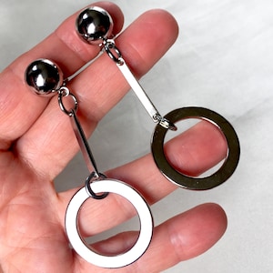 May include: A pair of silver earrings featuring a large circle and a smaller circle, both with a smooth, shiny finish. The earrings are suspended from a silver bar and a silver ball.