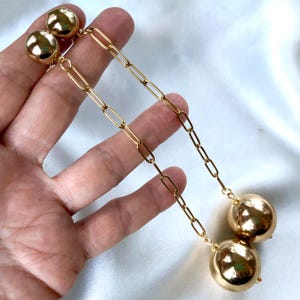 May include: Gold-coloured earrings with a large, round, polished ball at the bottom and a smaller ball at the top. The earrings are connected by a chain-link design. The jewellery is displayed against a white background.