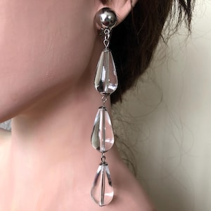 May include: Silver-toned dangle earrings featuring a large, round stud and three clear, teardrop-shaped pendants. The pendants are connected by delicate silver chains, creating a long, elegant design. The earrings are designed to be worn on the ear.