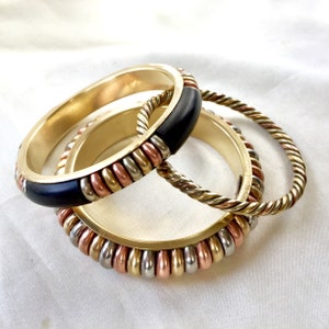 May include: Three gold bangles with black and copper accents. The bangles are stacked on top of each other. The top bangle has a black band with copper and silver beads. The middle bangle is a twisted gold rope design. The bottom bangle has a black band with copper and silver beads.