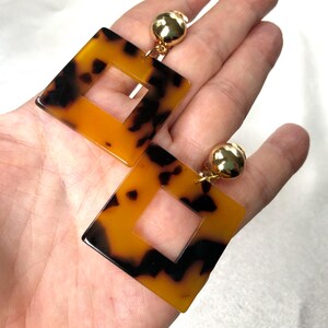 May include: A pair of gold tone clip-on earrings with a square-shaped tortoiseshell design. The earrings have a gold tone metal backing and a square-shaped hole in the centre.