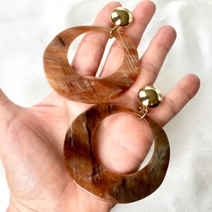 May include: A pair of large, brown, round hoop earrings with a gold-toned metal accent. The earrings have a marbled pattern.