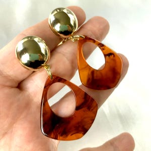 May include: A pair of gold tone clip-on earrings with large, brown, marbled, teardrop-shaped dangles. The dangles have a large, open circle in the center.