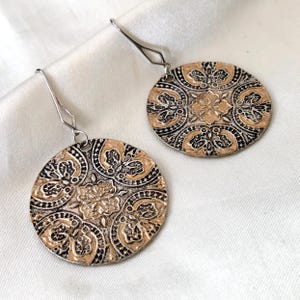 May include: A pair of silver-tone earrings with a circular, ornate design. The earrings feature a detailed pattern in shades of gold and black, with a floral motif at the center. The earrings are displayed on a white, silky surface.