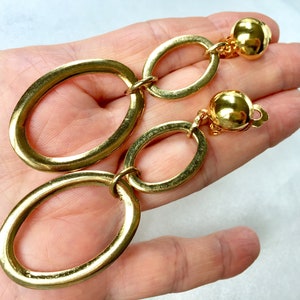 May include: A pair of gold-tone clip-on earrings featuring two oval-shaped links connected by a smaller oval link. The earrings have a textured finish and a round ball at the top.
