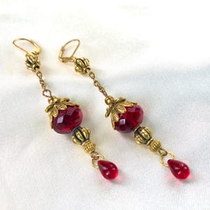 May include: A pair of long, dangling earrings with gold-toned metal accents and red faceted glass beads. The earrings have a teardrop-shaped bead at the bottom.