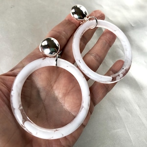 May include: A pair of large, white hoop earrings with a marbled design. The hoops are attached to silver-colored, round studs. The earrings are a modern accessory, suitable for various outfits.