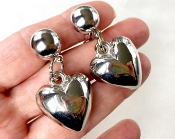 May include: A pair of silver clip-on earrings with heart-shaped charms. The charms are large and have a shiny, metallic finish.