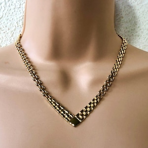 May include: A gold-coloured necklace with a V-shaped pendant. The necklace features a double-strand chain design that connects to a rectangular pendant. The jewellery is displayed on a mannequin.