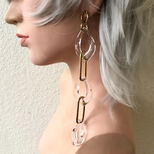 May include: A long drop earring featuring an alternating chain design. The earring consists of three clear, irregular, organic-shaped acrylic links connected by two smooth, elongated, gold-toned metal oval links in a vertical, dangling arrangement.