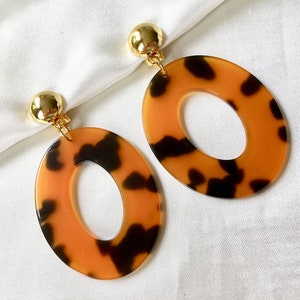 May include: A pair of oval-shaped clip-on earrings. The earrings feature a tortoiseshell pattern in shades of brown and orange. The clip-on tops are gold-colored and round.