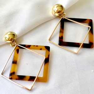 May include: A pair of geometric earrings with a gold-coloured stud and a square pendant. The pendant has a tortoiseshell pattern with a gold-coloured square frame. The earrings are displayed on a white surface.