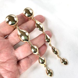 May include: A pair of long gold dangle earrings with a series of oval-shaped beads. The earrings are made of metal and have a shiny finish.