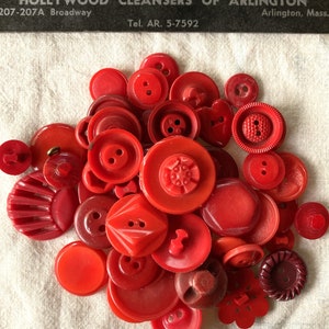 May include: A collection of red and burgundy plastic buttons of various shapes and sizes. The buttons are arranged in a pile on a white fabric surface.
