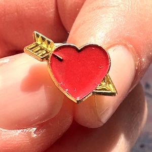 Pode incluir: Um alfinete dourado com um coração vermelho perfurado por uma flecha dourada. O coração é vermelho vibrante, e a flecha tem um design detalhado de penas. O alfinete é provavelmente um acessório com tema de Dia dos Namorados ou romântico.