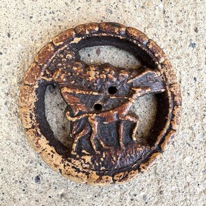 May include: A round, brown button with a deer and tree design. The button has two holes and a textured, decorative border. The deer is depicted in a running pose, and the button is made of a ceramic material.