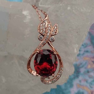 May include: A rose gold pendant necklace featuring a large red gemstone encircled by smaller clear gemstones. The pendant is shaped like a leaf with a swirling design.
