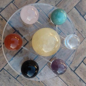 May include: Seven crystal spheres of various colors, including pink, green, orange, yellow, clear, black, and purple, are arranged in a circular pattern on a clear acrylic stand.
