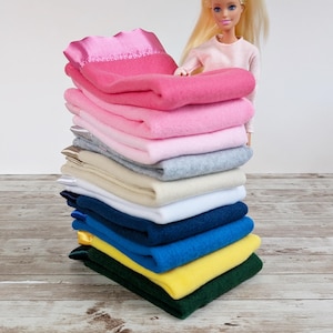 May include: A stack of folded blankets in various colors, including pink, gray, white, and blue. The blankets are neatly arranged, showcasing a range of hues. The top blanket is a vibrant pink with a satin trim. The blankets are stacked on a wooden surface.