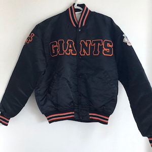 May include: Black satin baseball jacket with orange and grey striped trim. The jacket has a button closure and features the word "GIANTS" in orange embroidery on the front.
