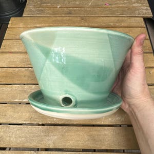 May include: A handcrafted, light green ceramic planter with a matching saucer. The planter has a unique, angular design and a drainage hole. The saucer has a white rim. The planter is designed for indoor or outdoor use.