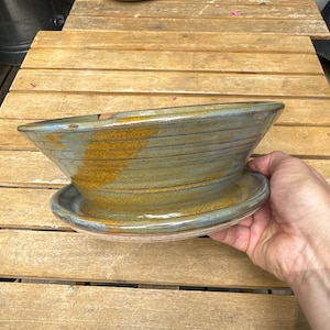 May include: A handcrafted ceramic planter with a matching saucer. The planter is a muted blue-green with streaks of gold and brown. The bowl-shaped planter is approximately 12 inches wide and 6 inches tall, perfect for succulents or small plants.