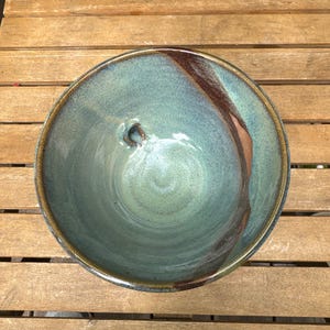 May include: A handmade ceramic bowl with a turquoise and brown glaze. The bowl has a circular shape with a slightly uneven rim. The interior is a blend of turquoise and green hues, with a brown accent.