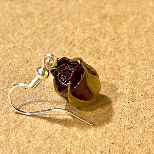 May include: A single silver earring with a dark red rose bud. The rose bud has green and brown leaves. The earring hook is silver and has a small ball at the top.