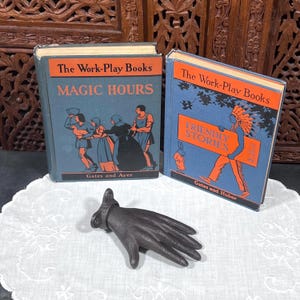 May include: Two vintage children's books titled "Magic Hours" and "Friendly Stories" with blue covers and orange text. A black cast iron hand-shaped object rests on a white lace cloth. The books are from "The Work-Play Books" series.