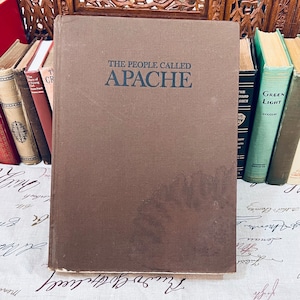 May include: A brown hardcover book titled "THE PEOPLE CALLED APACHE" in dark gray lettering. The book is surrounded by other vintage books with various colors and titles. The book's cover has a slightly worn appearance, suggesting its age.