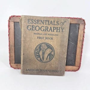 May include: A vintage hardcover textbook titled Essentials of Geography First Book by Brigham and McFarlane, published by American Book Company, featuring a circular illustration, positioned in front of a rectangular wooden-framed slate chalkboard.