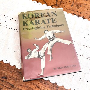 May include: A vintage book titled "KOREAN KARATE Free Fighting Techniques" with a cover illustration of two figures in white karate uniforms. The book is on a wooden surface with a white lace doily.