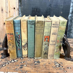 May include: A collection of eight vintage hardcover books held between two dark metal hand-shaped bookends. The spines feature various titles including The Boy Scouts in the Big Timber, Sturdy and Strong, Toby Tyler, and The Newsboy Partners.