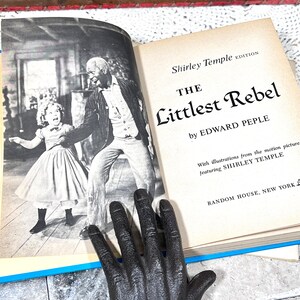 May include: An open book titled "The Littlest Rebel" by Edward Peple, a Shirley Temple edition. The book features a black and white photo of Shirley Temple and a man. A black hand is in the foreground.