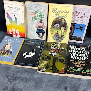 May include: A collection of vintage paperback books with colorful covers. Titles include "The Wild Duck," "Uncle Tom's Cabin," "Wuthering Heights," and "To Kill a Mockingbird." The books are arranged on a dark surface, showcasing their aged appearance.