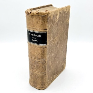 May include: An antique book with a worn, brown leather cover. The spine features a black label with the title "PLAIN FACTS" and the name "KELLOGG." The edges of the book show signs of age and wear, adding to its vintage appeal.