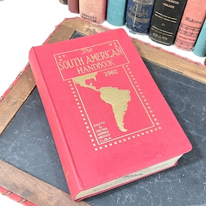 May include: A vintage, red hardcover book titled "The South American Handbook 1962." The cover features a gold map of South America and the text "South & Central America Mexico Cuba." Other books are visible in the background.