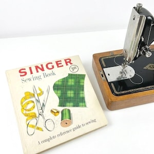 May include: A vintage Singer sewing machine with a black and silver finish, set on a wooden base. Beside it is a Singer Sewing Book with scissors, a measuring tape, and thread spools. The book cover features the text "Singer Sewing Book".