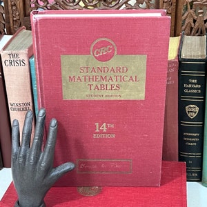 May include: A red textbook titled "Standard Mathematical Tables" with gold lettering and a CRC logo. The book is the 14th edition, student edition. Other books are visible in the background, along with a dark gray hand sculpture.