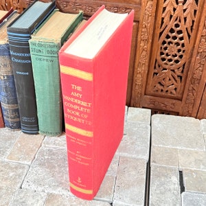 May include: A red hardcover book titled "The Amy Vanderbilt Complete Book of Etiquette" with gold accents. The book is standing upright next to other vintage books with various colors and titles. The book is on a stone surface.