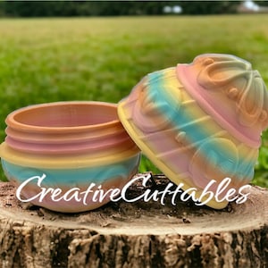 May include: A colorful, pastel-colored, round, ceramic container with a lid. The container has a swirl pattern and the lid has a crown design. The container is sitting on a tree stump. The text "CreativeCuttables" is visible on the container.