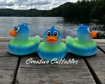 May include: Three blue and green rubber duckies with orange beaks and white eyes are sitting on a wooden dock. The duckies are facing forward and the dock is in front of a lake with green hills in the background.