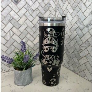 May include: Black stainless steel tumbler with a silver design of a woman with a soccer ball in her hair, sunglasses, and the words "Soccer Mom". The tumbler has a black lid with a silver ring.