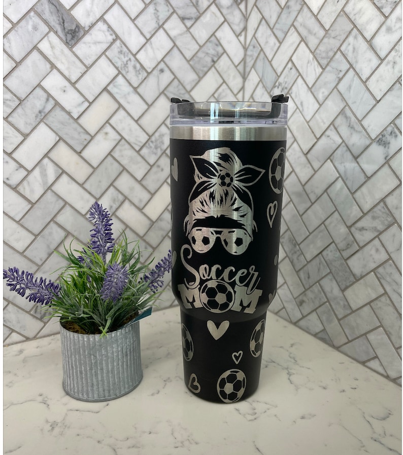 May include: Black stainless steel tumbler with a silver design of a woman with a soccer ball in her hair, sunglasses, and the words "Soccer Mom". The tumbler has a black lid with a silver ring.