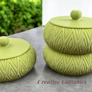 May include: Two green ceramic storage containers with lids. The containers have a geometric pattern and are stacked on top of each other. The containers are on a gray surface. The text "Creative Cuttables" is visible in the bottom right corner of the image.