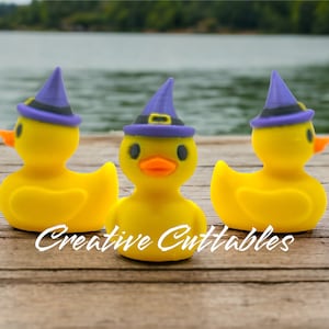 May include: Three yellow rubber duckies wearing purple witch hats with black bands. The ducks have orange beaks and black eyes. The text "Creative Cuttables" is written in white script across the bottom of the image. The ducks are on a wooden dock.