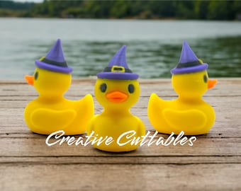 May include: Three yellow rubber duckies wearing purple witch hats with black bands. The ducks have orange beaks and black eyes. The text "Creative Cuttables" is written in white script across the bottom of the image. The ducks are on a wooden dock.