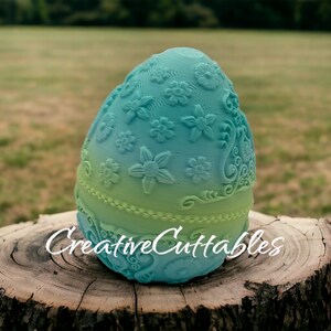 May include: A teal and green ombre egg-shaped mold with a floral design. The mold is sitting on a tree stump. The text "Creative Cuttables" is visible on the mold.